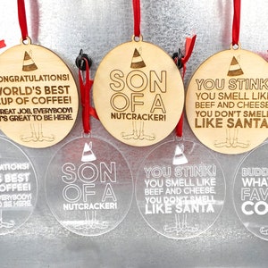 May include: A collection of round Christmas ornaments with humorous quotes and elf illustrations. Some are clear acrylic, others are wood. Each ornament has a red ribbon for hanging. The text includes phrases like "Son of a Nutcracker!" and "You Stink!"