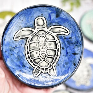 May include: A blue ceramic coaster with a silver turtle design. The coaster has a speckled blue glaze and a white rim.