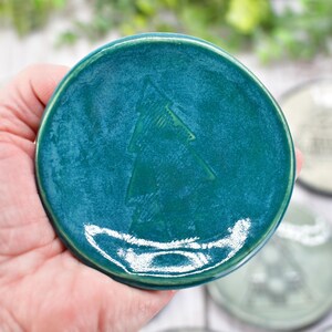May include: A teal ceramic coaster with a raised Christmas tree design. The coaster is round and has a glossy finish. Other coasters with different designs are visible in the background.