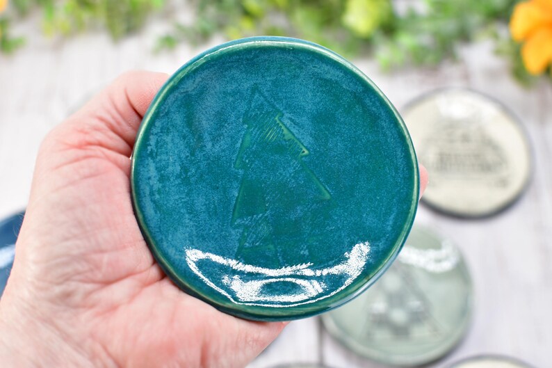 May include: A teal ceramic coaster with a raised Christmas tree design. The coaster is round and has a glossy finish. Other coasters with different designs are visible in the background.
