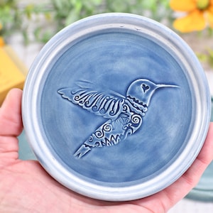 May include: A blue ceramic coaster with a raised hummingbird design. The hummingbird has intricate wing and body details, and a heart shape on its head. The coaster has a light blue rim and is held in a hand.