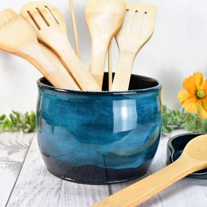 May include: A teal ceramic utensil holder with a black rim, filled with wooden kitchen utensils. A matching teal ceramic spoon rest with a wooden spoon is on the table.