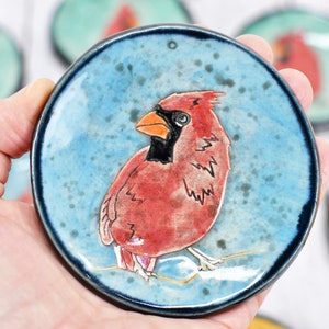 May include: A ceramic dish with a blue glaze and a hand-painted cardinal bird. The cardinal is red with a black head and a yellow beak. The dish is about 4 inches in diameter.