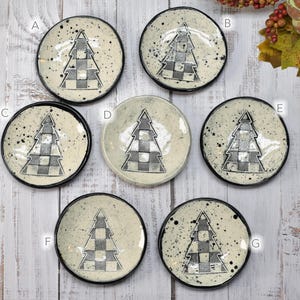 May include: A collection of seven small, round ceramic dishes. Each dish features a black and white checkered Christmas tree design in the center. The dishes have a speckled, off-white background and black rims.