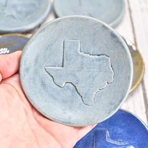 May include: A round, slate-blue ceramic coaster featuring a debossed outline of the state of Texas in the center. The surface has a matte, textured finish with subtle variations in color, and the edges are slightly raised to form a shallow dish.