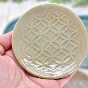 May include: A small, round, olive-green ceramic dish featuring a raised, repeating geometric pattern of interlocking circles and diamonds across its surface. The dish has a smooth, glossy finish and a slightly irregular, hand-formed circular edge.