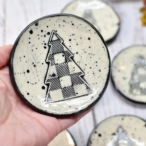 May include: A set of handmade ceramic coasters with a black and white buffalo plaid Christmas tree design. The coasters have a speckled cream background and a black rim. The image shows a hand holding one coaster, with several others in the background.