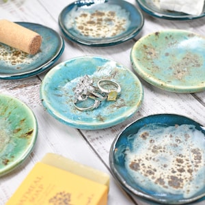 May include: A collection of small, round ceramic dishes featuring a glossy glaze in shades of teal, turquoise, and seafoam green with speckled brown accents. Some dishes hold items like a cork, metal rings, a silver spoon, and white soap.