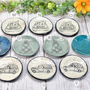 May include: A collection of small, round ceramic dishes with various Christmas-themed designs. Some feature a truck carrying a Christmas tree, others have a tree design, and some say "Merry Christmas". The dishes are in shades of blue, green, and white.