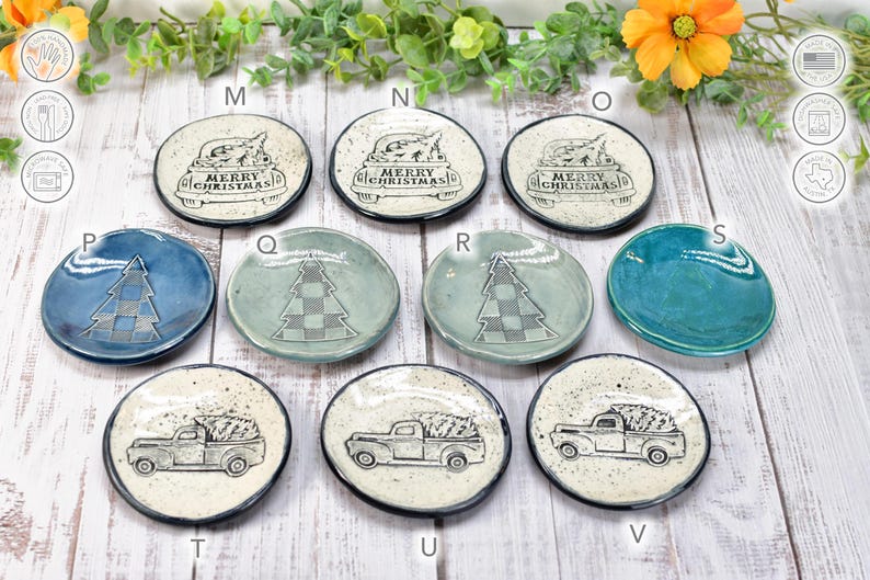 May include: A collection of small, round ceramic dishes with various Christmas-themed designs. Some feature a truck carrying a Christmas tree, others have a tree design, and some say "Merry Christmas". The dishes are in shades of blue, green, and white.