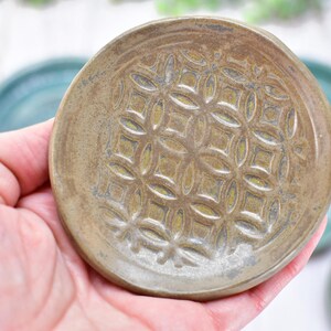 May include: A small, round, tan-colored ceramic dish featuring a raised, repeating geometric pattern of interlocking circles and squares on its surface. The dish has a smooth, glazed finish with subtle variations in tone across the textured design.