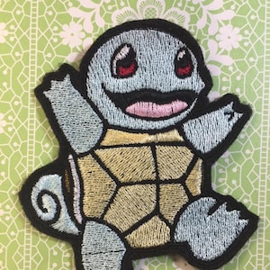 May include: A blue and yellow embroidered patch of a cartoon turtle with a big smile. The turtle has red eyes and a pink tongue. The patch is on a green and white background.
