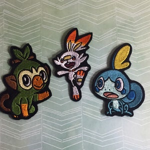 May include: Three embroidered patches featuring Pokemon characters: Grookey, Scorbunny, and Sobble. Grookey is green with brown accents, Scorbunny is orange and white, and Sobble is blue with yellow accents.