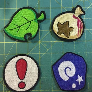 May include: Four embroidered patches featuring popular video game characters. The patches are green, beige, white, and blue. The first patch is a green leaf with a bite taken out of it. The second patch is a beige bag with a brown star and a red line. The third patch is a white circle with a red exclamation point. The fourth patch is a blue pentagon with a white spiral and a white star.