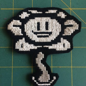 May include: Black and white embroidered patch of a smiling character with a flower-like head and a stem. The character has a pixelated appearance.