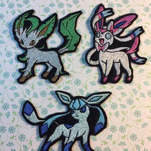 May include: Three embroidered iron-on patches featuring different Pokemon characters. The patches are on a white background with a floral pattern. The Pokemon characters are Eevee, Espeon, and Leafeon.