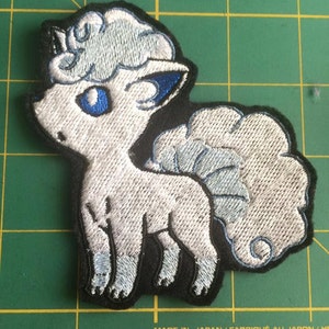 May include: White and blue embroidered patch of a Pokemon character, a fluffy, white, dog-like creature with a blue tail and blue eyes. The patch is on a green grid background.