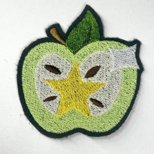 May include: An embroidered patch shaped like a sliced green apple. The design features a light green outer skin, a white interior flesh, and a central yellow star shape where the core would be. Four brown seeds are visible, with a green leaf on top.