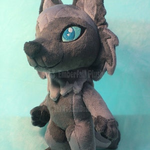 May include: Gray plush wolf with blue eyes. The wolf is standing on a light blue surface. The text "Emberfall Plus" is visible on the wolf's chest.