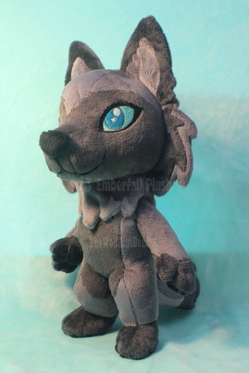 May include: Gray plush wolf with blue eyes. The wolf is standing on a light blue surface. The text "Emberfall Plus" is visible on the wolf's chest.
