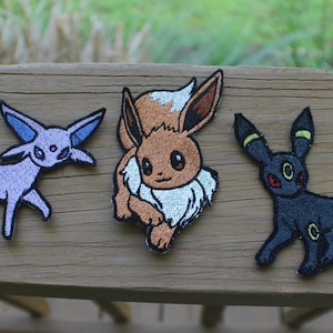 May include: Three embroidered iron-on patches featuring Pokemon characters: Eevee, Espeon, and Umbreon. The patches are on a wooden surface.