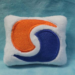 May include: A white plush pillow with an embroidered orange and blue swirl design. The swirl design resembles the logo of a popular laundry detergent brand.