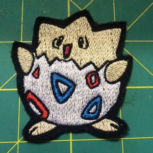 May include: Embroidered patch of a cartoon Pokemon character, a light brown and white Eevee, with a blue and red egg pattern. The Eevee is smiling with its tongue sticking out.