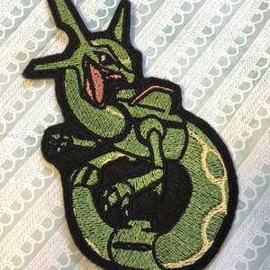 May include: Green and black embroidered patch of a Pokemon character. The character is a green, reptilian creature with a long, pointed tail and a large, pointed head. The character is curled up in a ball, with its tail wrapped around its body.