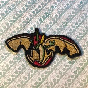 May include: A black, red, and green embroidered patch of a flying dinosaur with outstretched wings.