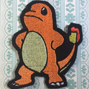 May include: An orange and black embroidered patch of a Pokemon character, Charmander, with a flame tail.