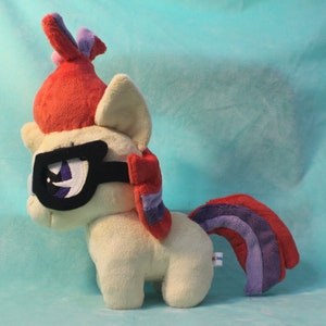 May include: A plush toy of a pony with a red and purple mane and tail. The pony is wearing black glasses and has a white body. The pony is standing on a light blue surface.