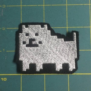 May include: A white embroidered patch of a pixelated dog on a black background. The patch is about 2 inches wide and 1.5 inches tall.
