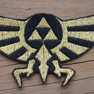 May include: A gold and black embroidered patch featuring the Triforce symbol from the Legend of Zelda video game series. The Triforce is surrounded by a stylized bird's wings.
