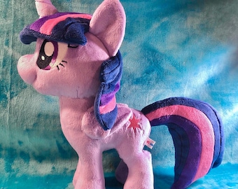 May include: A plush toy of a purple pony with a horn, blue and purple mane and tail, and a red star on its flank. The pony has large eyes and is standing on a blue surface. The text "@EmberfallPlush" is at the bottom.
