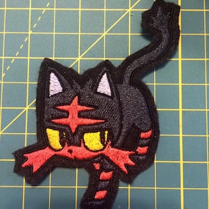 May include: Black and red embroidered patch of a cartoon cat with yellow eyes. The cat is in a crouching position with its tail curled up.