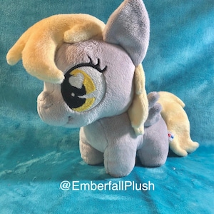 May include: A plush toy pony in shades of gray and yellow, with large, detailed eyes. The pony has a fluffy mane and tail, and the text "@EmberfallPlush" is visible.