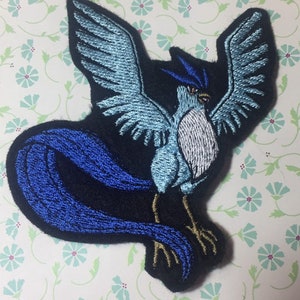 Peut inclure: Écusson brodé représentant un oiseau stylisé aux ailes déployées. L'oiseau présente des détails bleu clair et blancs, avec un contour et une queue bleu foncé. L'écusson est sur un fond noir et a des pieds dorés.