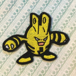 May include: Yellow and black embroidered iron-on patch of a cartoon character with a striped body and a wide grin.