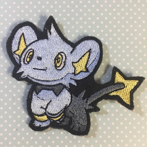 May include: An embroidered iron-on patch of a blue and grey Pokemon character with yellow accents. The character has large ears and a long tail.