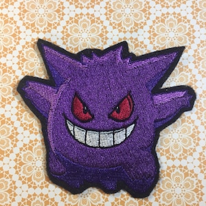 May include: Purple and black embroidered patch of a cartoon ghost with a wide, toothy grin and red eyes.