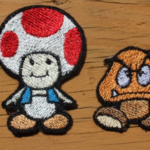 May include: Two embroidered patches featuring characters from the Super Mario Bros. video game series. The first patch is a white mushroom with a red spot on top, and a smiling face. The second patch is a brown mushroom with a frowning face and sharp teeth.