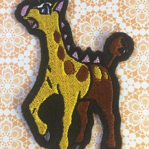 May include: Embroidered iron-on patch of a yellow and brown giraffe with purple accents. The giraffe has a long neck and a long tail. The patch is perfect for adding a touch of whimsy to clothing, bags, or other accessories.