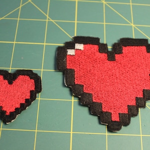 May include: Two red pixelated heart-shaped patches with black outlines. The larger heart is about 3 inches wide and the smaller heart is about 1.5 inches wide.