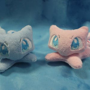 May include: Two plush toys, one blue and one pink, with embroidered eyes and a cartoonish appearance. The toys are lying on a blue fuzzy surface.