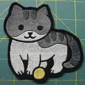 May include: Embroidered iron-on patch of a gray and white cat playing with a yellow ball.