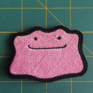 May include: Pink embroidered patch of a Pokemon character with a black outline. The character has a simple smiling face.
