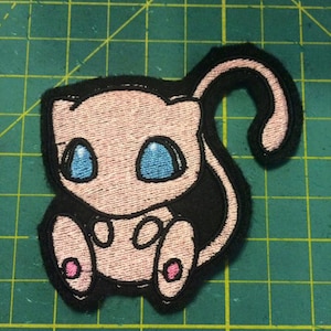 May include: Pink and black embroidered iron-on patch of a cartoon Pokemon character, Mew, with blue eyes and pink cheeks.