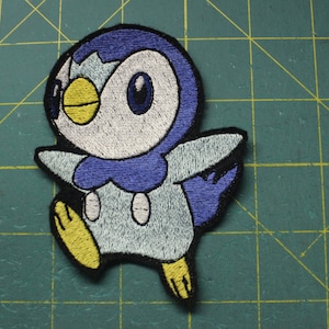May include: Embroidered patch of a blue and white penguin with yellow accents. The penguin is standing on its feet with its wings spread out.