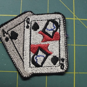 May include: A close-up of a white and gray embroidered patch featuring two playing cards. The cards are both spades, one is a jack and the other is a queen. The cards are partially overlapping.
