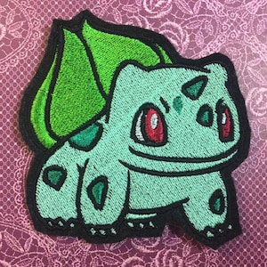 May include: Green and blue embroidered iron-on patch of a cartoon Pokemon character, Bulbasaur. The patch has black outlines and red eyes.